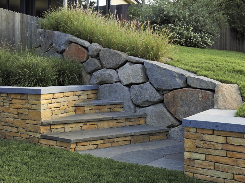 A sloped yard terraced with a wall of large fitted boulders above a stacked tan stone wall with blue-grey caps and stone steps.