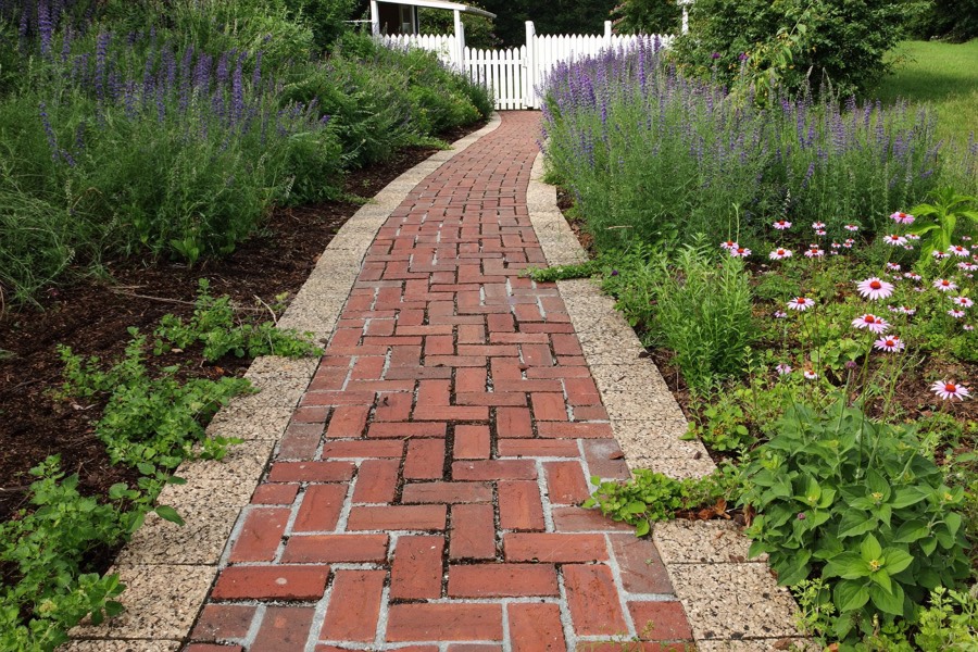 A red brick walkway with a pale edge running between flowering borders toward a white picket gate.