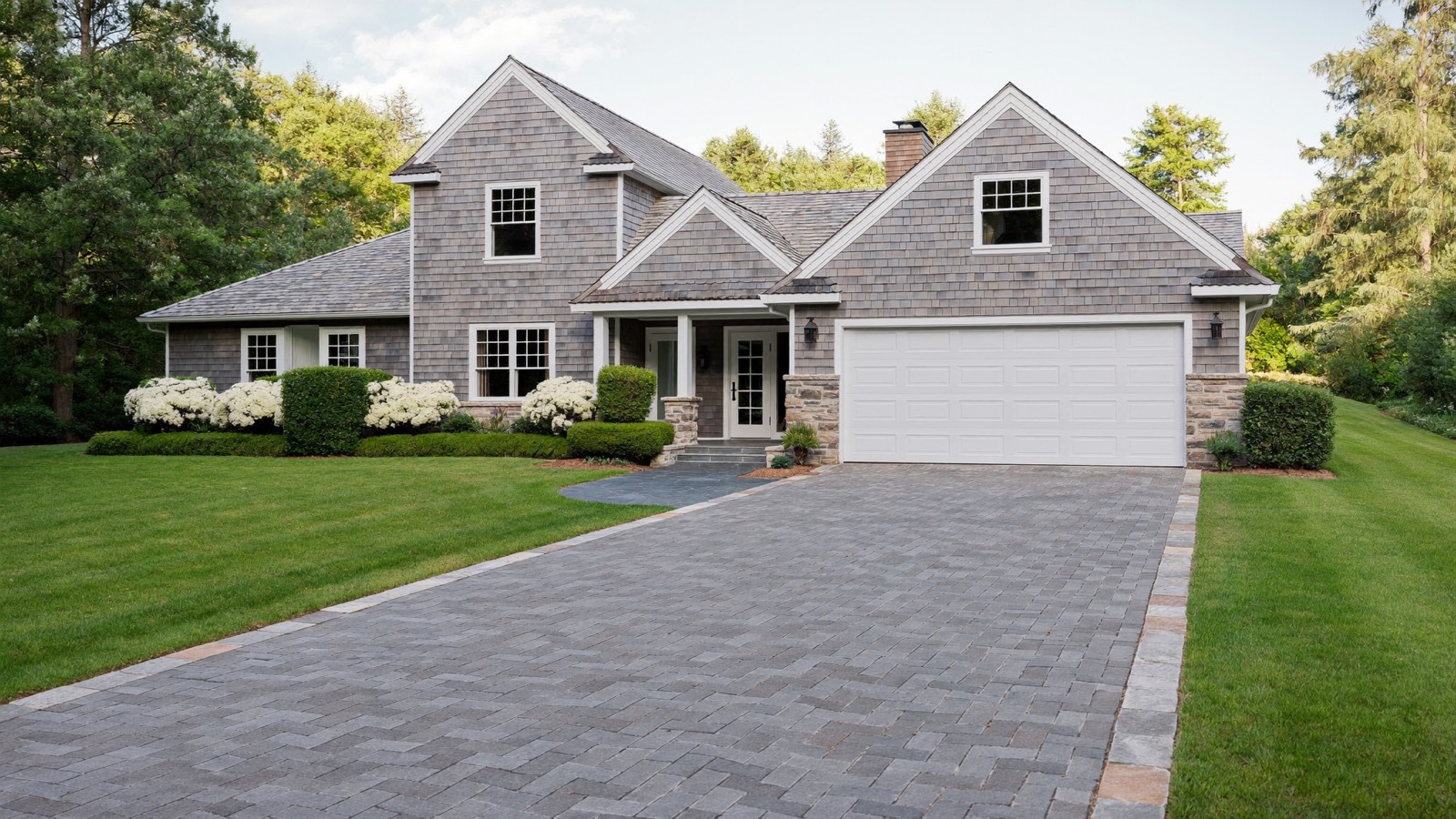 A grey shingled house with a white garage door, a herringbone paver driveway with lighter block edging, and a dark stone path to the porch.