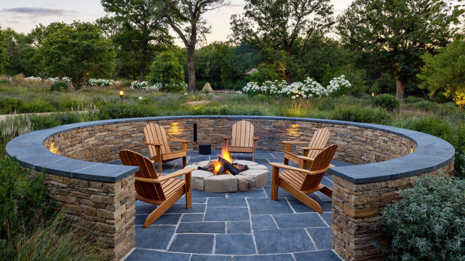 A round sunken seating area at dusk with a fire pit, four wooden Adirondack chairs and a curved stacked-stone wall with blue-grey caps.