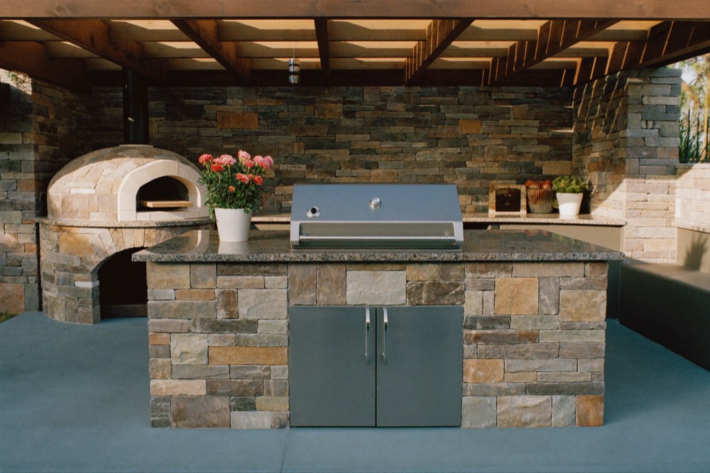 An outdoor kitchen under a wooden pergola: a stone-clad island with a stainless grill and a speckled stone counter, beside a domed stone pizza oven.