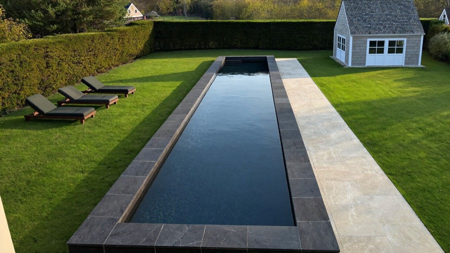 A long narrow pool with dark stone coping set into a lawn, a pale stone walk along one side and a small shingled pool house.