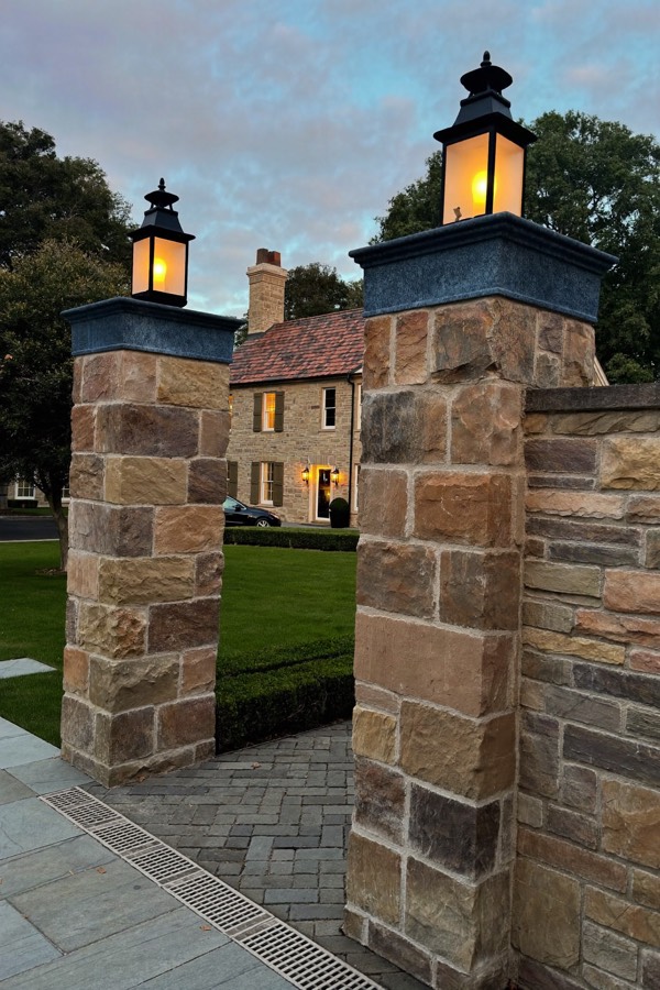 Two stone-clad pillars with dark caps and glowing lanterns at the entrance to a paver drive at dusk.