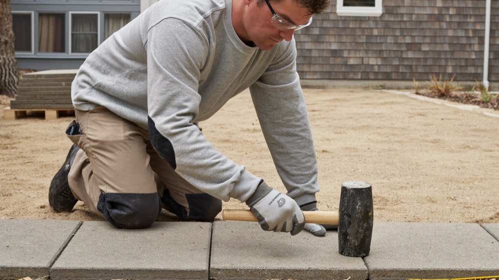 A man in safety glasses and knee pads kneels beside a row of grey pavers, tapping one into place with a rubber mallet, with a smooth sand bed behind him.