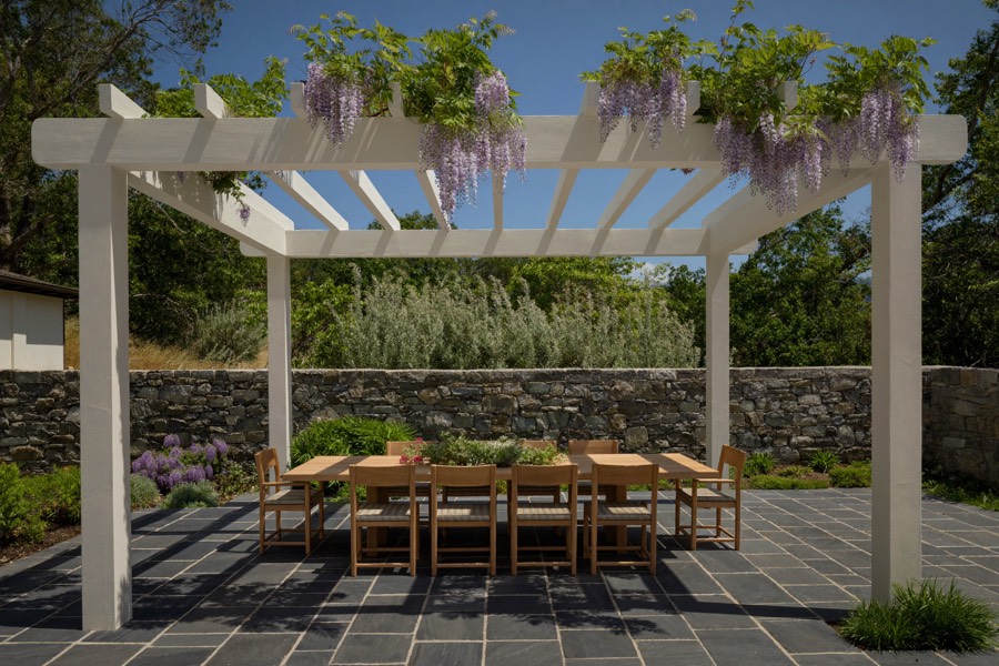 A long wooden dining table and chairs on dark stone paving under a white pergola hung with purple wisteria, in front of a stacked-stone wall.