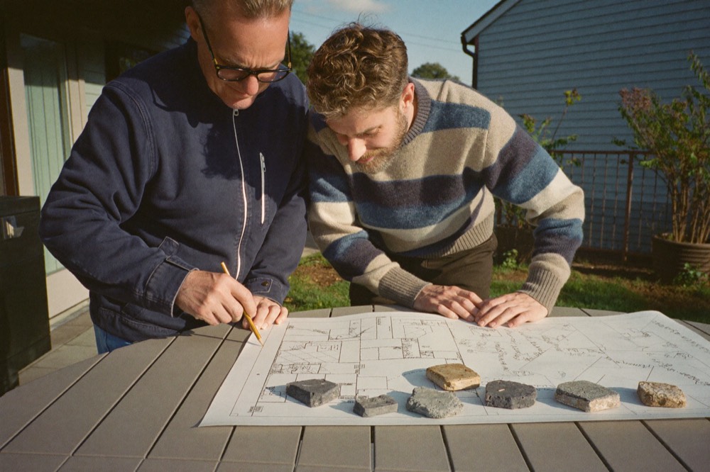 Two men lean over an outdoor table looking at a printed patio plan, one marking it with a pencil, with small stone samples laid along the bottom of the sheet.
