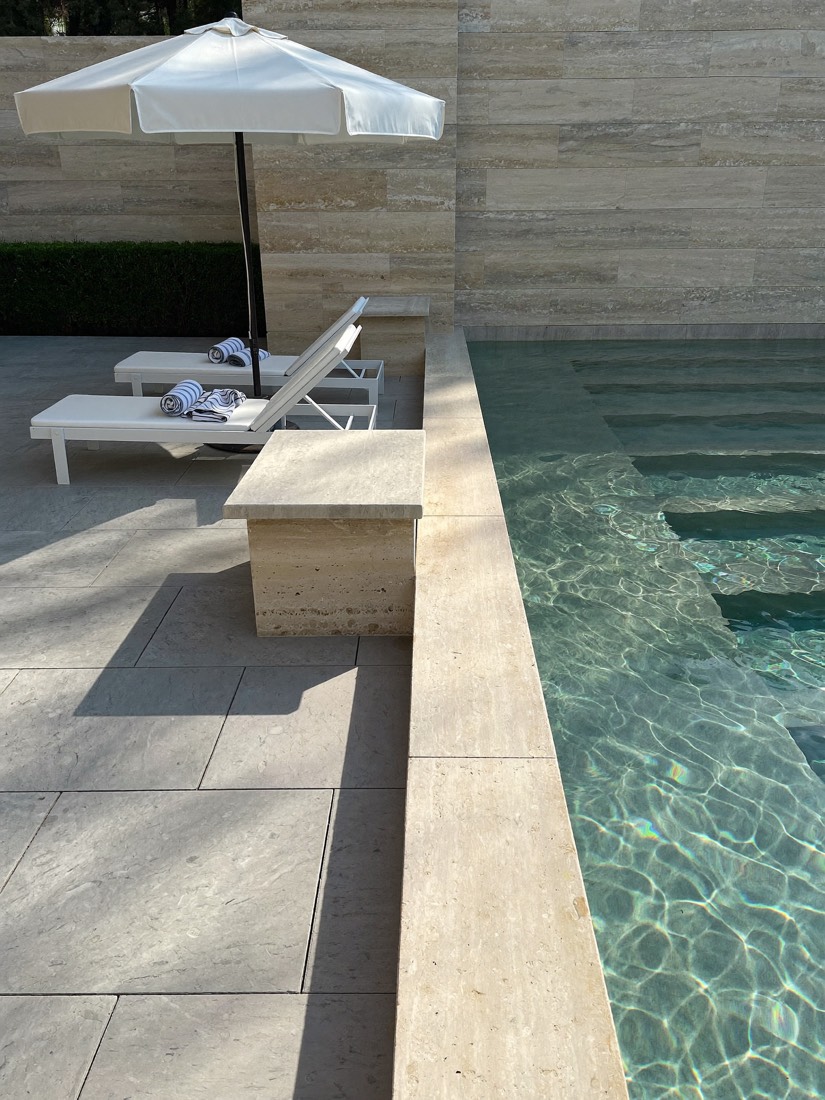 The edge of a pool with thick pale stone coping beside a light grey paved terrace, two white loungers and a white umbrella.