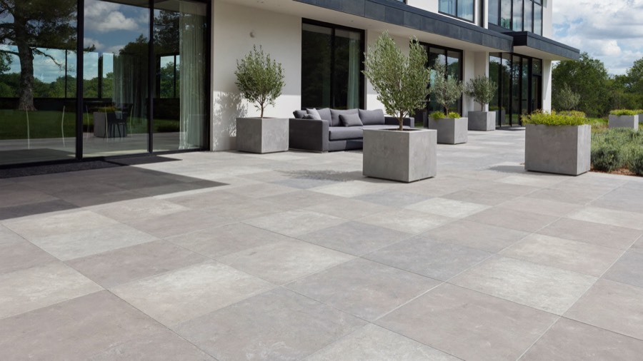 A wide terrace of large pale grey pavers beside a modern house with tall glass doors, with small trees in square planters.