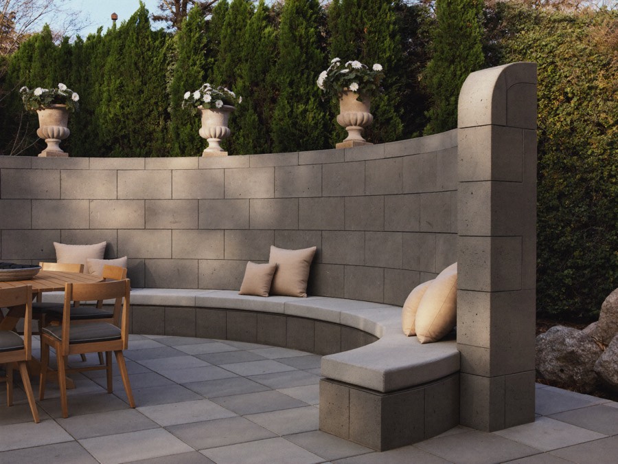 A curved grey block seat wall with a built-in bench and cushions around a paved patio, with stone urns along the top.