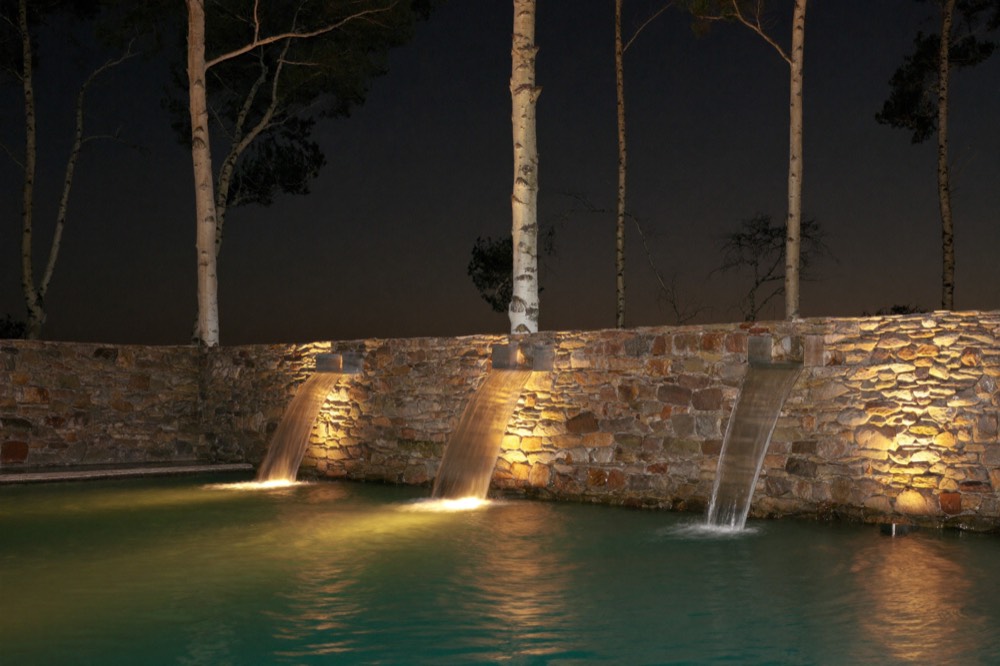 Three sheets of water falling from a lit stone wall into a pool at night, with white-barked trees behind.
