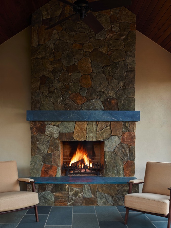 A tall stone fireplace with a blue-grey mantel and hearth, a wood fire burning and two armchairs facing it.